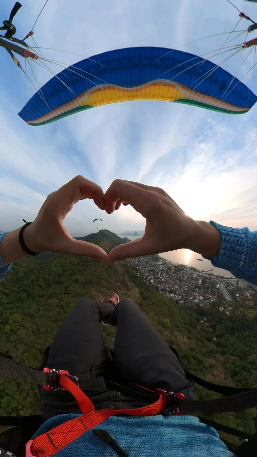 Passageiro fazendo coração com as mãos durante voo de parapente ao pôr do sol em Niterói