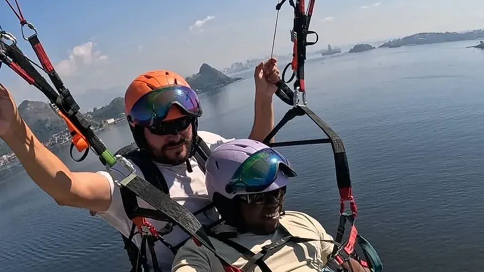Piloto e passageira de braços abertos em voo duplo de parapente sobre a Baía de Guanabara, com o Pão de Açúcar ao fundo